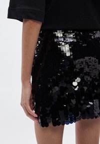 Czarna mini spódnica z cekinami w kształcie okrągłych, błyszczących dysków, noszona z luźną czarną bluzką. Charakteryzuje się prostym dołem i gładką fakturą.