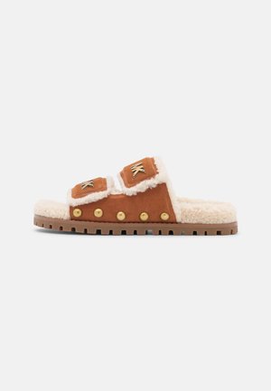 Sandale à glissière en daim marron avec doublure en shearling crème, clous dorés sur les côtés, semelle épaisse à crampons et deux sangles réglables avec des logos MK dorés.