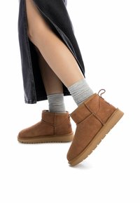 Botas de ante marrón hasta el tobillo con suela de goma plana, con una costura lateral y una pequeña lengüeta, combinadas con calcetines grises acanalados y una falda oscura.