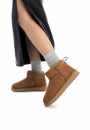 Botas de ante marrón hasta el tobillo con suela de goma plana, con una costura lateral y una pequeña lengüeta, combinadas con calcetines grises acanalados y una falda oscura.