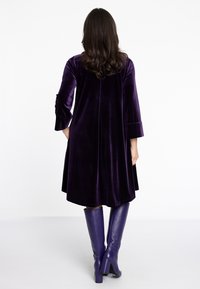 Lila Samtkleid mit Dreiviertelärmeln, ausgestelltem Schnitt und mittellanger Saumlänge, kombiniert mit knielangen lila Lederstiefeln.