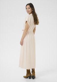 Robe maxi en tissu beige doux avec des manches courtes, détail de taille noué et texture subtile. Assortie à des bottes en daim marron à la cheville.