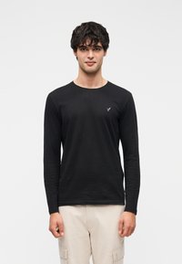 Tricou negru cu mâneci lungi, cu gât rotund și un mic logo pe piept, realizat dintr-un material moale, purtat cu pantaloni cargo de culoare deschisă.