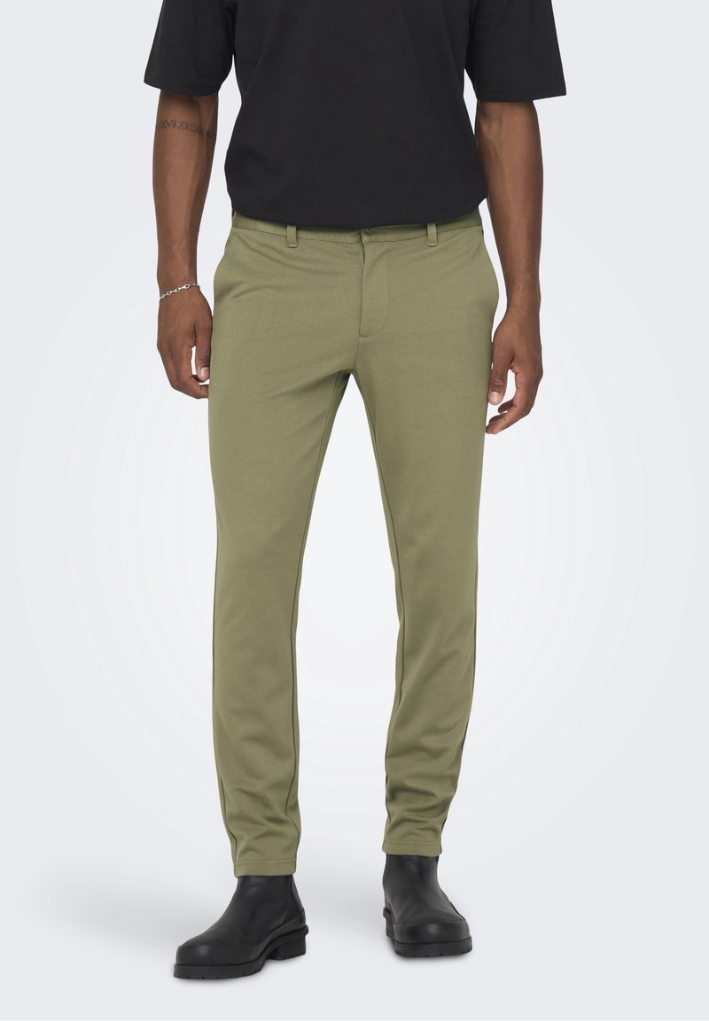 Pantalon vert kaki en tissu stretch, avec une coupe slim, devant plat, passants pour ceinture et coutures latérales discrètes. Porté avec des bottes noires.