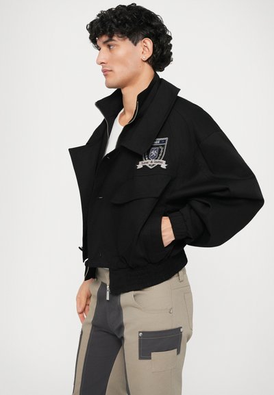 Zwarte cropped jas met een hoge kraag, twee voorzakken en een geborduurd embleem. Draagt met beige en grijze cargobroek.