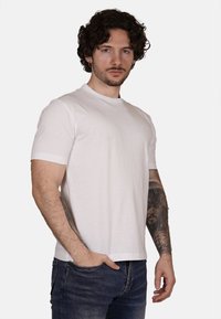 Maglietta bianca con collo rotondo realizzata in cotone, con maniche corte e una vestibilità rilassata. Indossata con jeans in denim scuro, che mostrano tatuaggi sulle braccia.