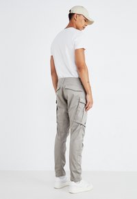 Grå cargo-bukser med flere sidelommer, smal passform og bomullsmateriale. Kombinert med en hvit T-skjorte og hvite joggesko, iført en beige caps.