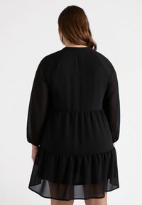 Vestido negro con mangas largas y transparentes y un dobladillo de volantes en capas. Presenta un ajuste relajado y una cinturilla elástica, hecho de una tela ligera.