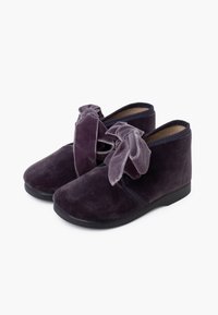 Botas de tobillo de terciopelo púrpura con punta redondeada, paneles laterales elásticos y un gran lazo en la parte delantera; suelas de goma negras para mayor agarre.