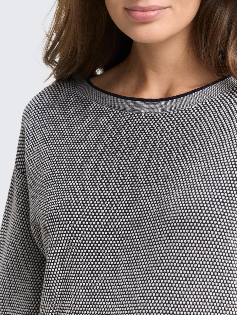 Kobieta nosząca czarną i białą teksturowaną dzianinową bluzkę z okrągłym dekoltem, ukazującą szyję, podbródek i część długich brązowych włosów.