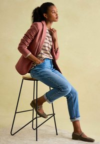 Blazer aus pinkem Stoff über einem gestreiften Hemd, kombiniert mit hellblauen, umgeschlagenen Jeans und braunen Ballerinas mit einer Schnalle. Sitzend auf einem schwarzen Hocker.
