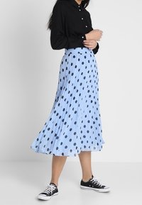 Gonna plissettata azzurro chiaro con pois neri, abbinata a una camicia nera con bottoni e sneakers nere. La gonna presenta un elastico in vita.