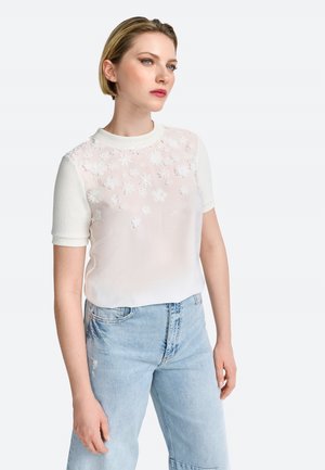 Witte top met korte mouwen en bloemenborduursel over de voorkant, semi-transparante voorkant en effen kleur mouwen. G gedragen met lichtblauwe jeans.