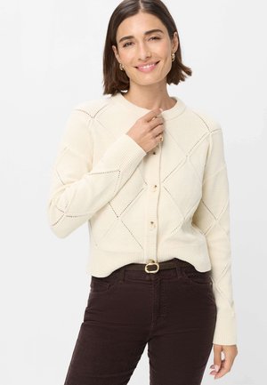 Femme aux cheveux courts bruns, souriante, portant un cardigan en tricot crème et un pantalon marron foncé avec une ceinture noire sur un fond uni.