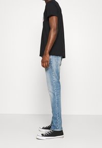 Camiseta de algodón negra, vaqueros rectos azul claro y zapatillas de lona negras con suelas de goma blancas, vistos de perfil.