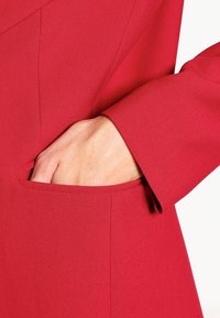 Röd skräddarsydd jacka med en slät tygstruktur, med en sidficka och en figursydd stil. Ärmen sträcker sig precis ovanför handleden.