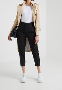 Trench coat beige sopra un top bianco a costine, pantaloni neri cropped e sneakers bianche. Borsa nera con tracolla.