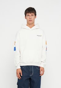 Sweatshirt branco com capuz e bolso frontal, apresentando um logótipo preto e detalhes coloridos nas mangas. Usado com calças listradas escuras.