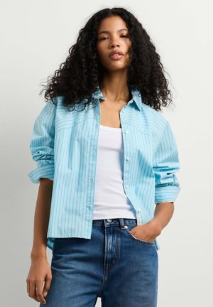 Ung kvinde med krøllet hår iført en lyseblå stribet skjorte over en hvid tanktop og blå jeans, med den ene hånd i lommen.