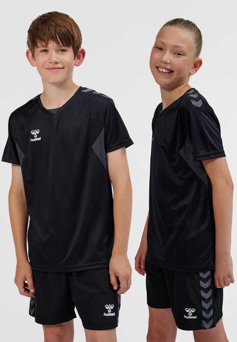 Twee kinderen in zwarte sportkleding met hummel-logo's, zij aan zij staand, glimlachend tegen een effen lichte achtergrond.