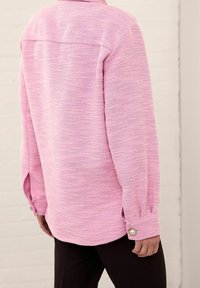 Person wearing a textured pink long-sleeve shirt with pearl-buttoned cuffs and dark pants, standing against a light brick wall.