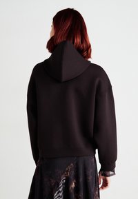 Svart, kort hoodie med stor hette, avslappet passform og ribbestrikkede mansjetter. Teksturen ser myk ut, og er kombinert med et mønstrete skjørt.