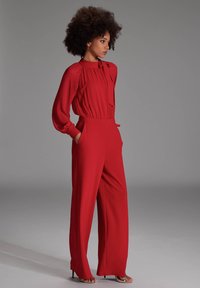 Rode jumpsuit met lange mouwen, geplooid ontwerp, strikdetail bij de hals, wijde pijpen en zakken. Glad textuur van de stof.