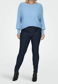 Kvinna som bär en ljusblå ribbad tröja, mörka skinnyjeans, svarta ankelstövlar och ett guldhalsband, stående mot en vit bakgrund.