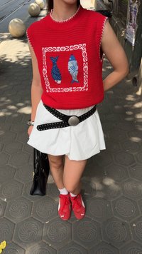 Top sin mangas de punto rojo con diseños de peces; falda blanca con pliegues; cinturón negro con tachuelas; zapatillas rojas; bolso negro; fondo de pavimento texturizado.