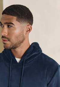 Felpa con cappuccio in corda blu navy, dotata di un cordino di regolazione, caratterizzata da un tessuto texturizzato e un cappuccio ampio. Primo piano del profilo laterale di un uomo.