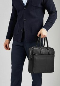Sort læder laptop taske med to håndtag, en lynlåslukning, udvendig lomme og tekstureret overflade. Båret af en person i en mørk cardigan.