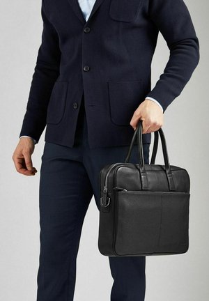 Sort læder laptop taske med to håndtag, en lynlåslukning, udvendig lomme og tekstureret overflade. Båret af en person i en mørk cardigan.