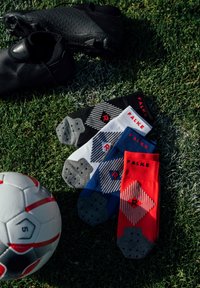 Botas de fútbol negras y cuatro pares de calcetines FALKE en negro, blanco, azul y rojo, con patrones geométricos y agarres de goma.