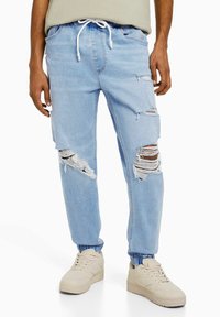 Ljusblå rivna jeans med elastiska muddar, som bärs med beige sneakers, visas på en person som står mot en vit bakgrund.