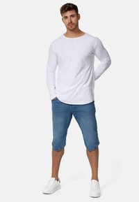 Camisa blanca de manga larga con un corte holgado, combinada con pantalones cortos de denim azul hasta la rodilla. Zapatillas blancas completan el atuendo.