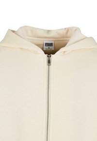 Sweat à capuche zippé couleur crème avec une fermeture éclair métallique et une étiquette de marque "Urban Classics" visible à l'intérieur du col, taille M.