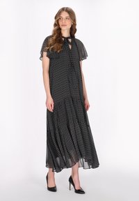 Maxi vestido preto estampado com decote amarrado, mangas curtas de babados e barra em camadas. Usado com saltos altos pretos. Tecido leve e fluido.