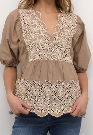 Femme portant un chemisier beige à manches bouffantes avec des détails en dentelle florale sur le col et l'ourlet, assorti à un jean bleu.