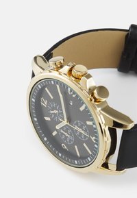 Montre chronographe dorée avec cadran noir, index métalliques, et bracelet en cuir. Équipée de poussoirs et d'accents texturés sur le boîtier.