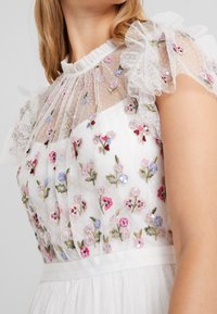 Valkoinen mekko, jossa on kuvioitu kirjailtu yläosa värikkäillä kukkakuoseilla, röyhelöiset läpikuultavat hihat ja satiinivyötärö.