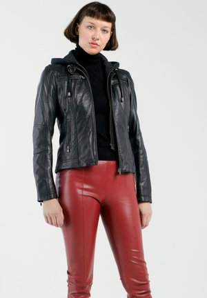 Vrouw draagt een zwarte leren jas over een zwarte coltrui en strakke rode leren broek, staand tegen een effen lichte achtergrond.