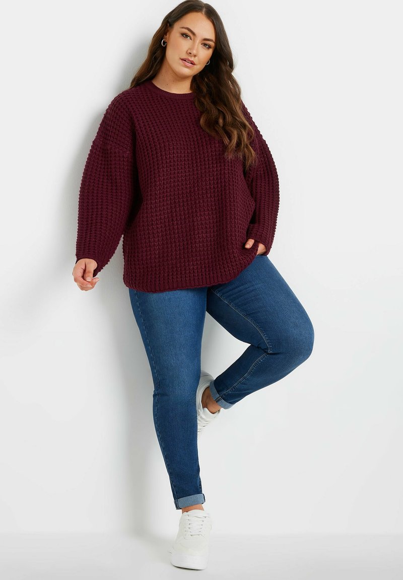 Bordowy sweter z dzianiny waflowej o luźnym kroju i długich rękawach, połączony z niebieskimi jeansami typu skinny i białymi sneakersami. Miękka tekstura, casualowy design.