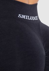 Zwarte sporttop met een hoge halslijn, een gladde textuur en het "SMILODOX" logo in het wit op de kraag. Strak passende pasvorm.