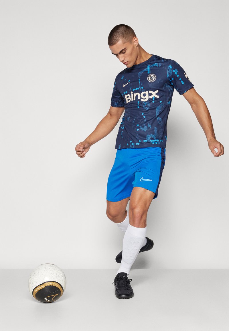 Maglietta da calcio Nike blu con motivo a spruzzi, abbinata a pantaloni corti blu e calze bianche. Scarpe da calcio nere. Una palla bianca nelle vicinanze.