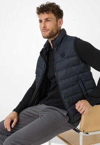  Gepolsterte, dunkelblaue Jeansweste mit hohem Kragen, Reißverschluss und schwarzen Akzenten. Getragen über einem schwarzen Langarmshirt, kombiniert mit grauen Hosen.