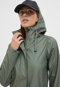 Green waterproof jacket with a hood, zip closure, and snap buttons. Features drawstring and rubber accents, paired with a dark cap.