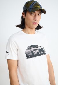 Biała koszulka bawełniana z czarną grafiką samochodu z przodu; dekoracyjna logo Puma na rękawie. Model nosi zieloną czapkę w wzory.