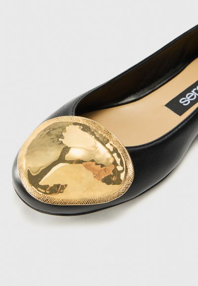 Svart läderballerina med en stor, oval guldmetalltåhuvud med texturerad kant och slät yta.