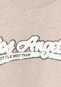 Tissu tricoté beige avec texte brodé blanc flou indiquant "Los Angeles" et texte plus petit en dessous indiquant "THE BEST TEAM."