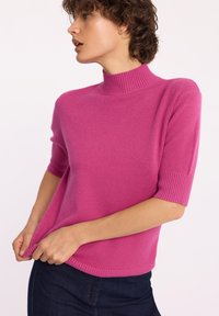 Różowy sweter z krótkim rękawem i wysokim, prążkowanym dekoltem, o miękkiej fakturze i dyskretnych prążkowanych detalach na rękawach i dolnym wykończeniu. Zestawiony z ciemnymi dżinsami.